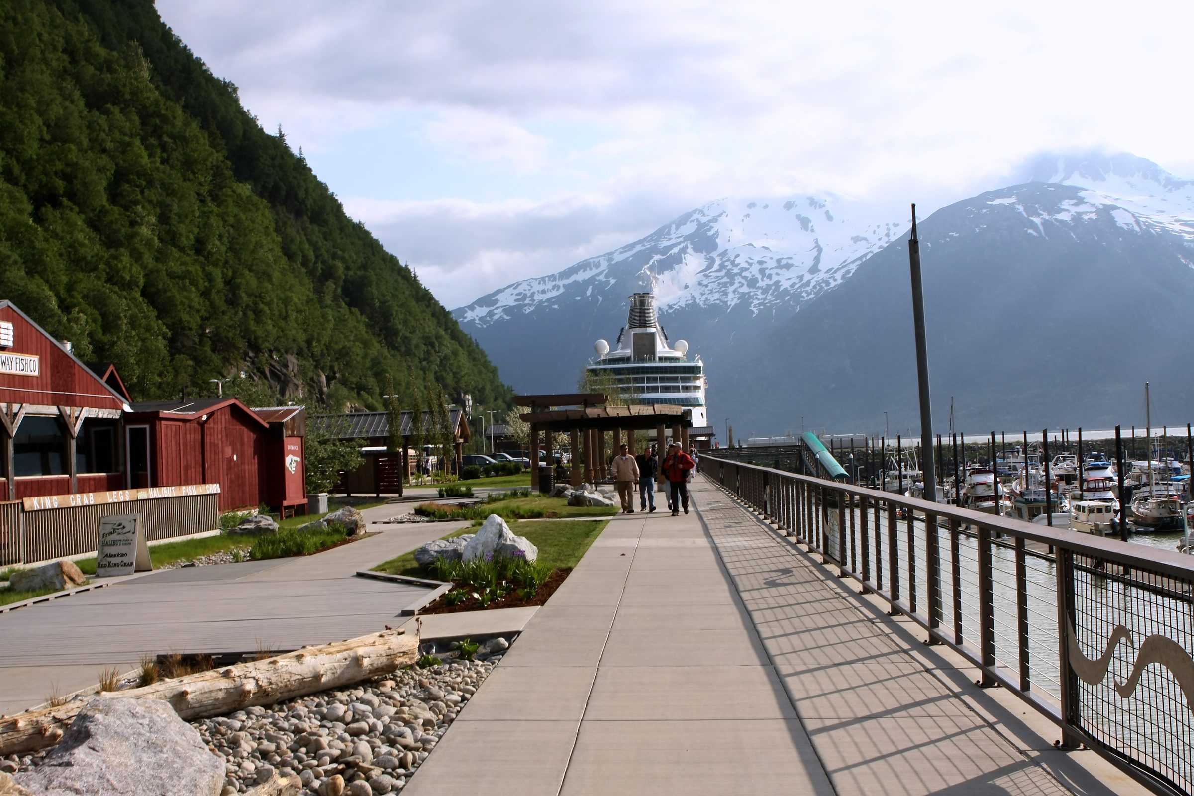 Wooden boardwalk with red harbor buildings and snowy peaks beyond.