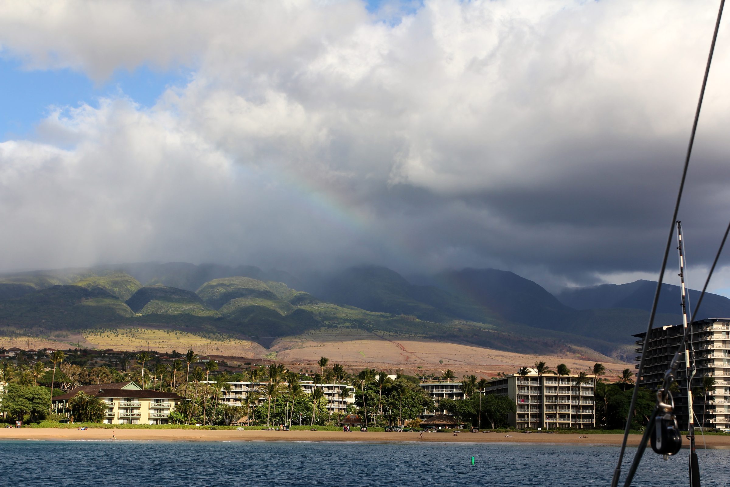 Coastal resort town with palms under cloud-wrapped hills and a faint rainbow.