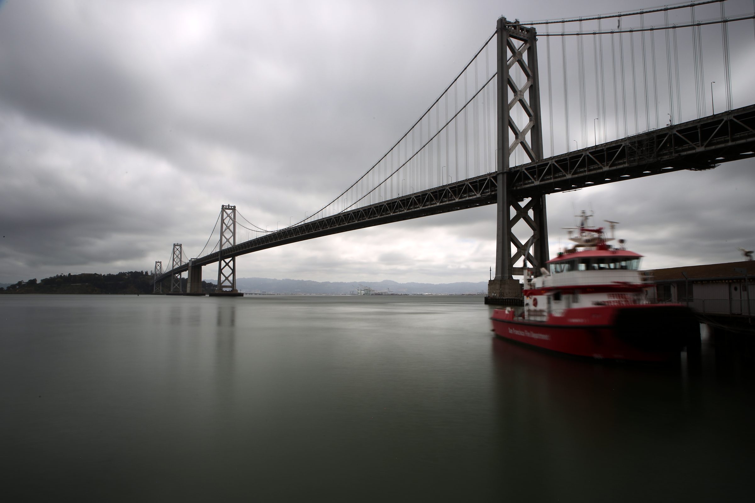 Low-angle Bay Bridge with a red fireboat under grey sky.