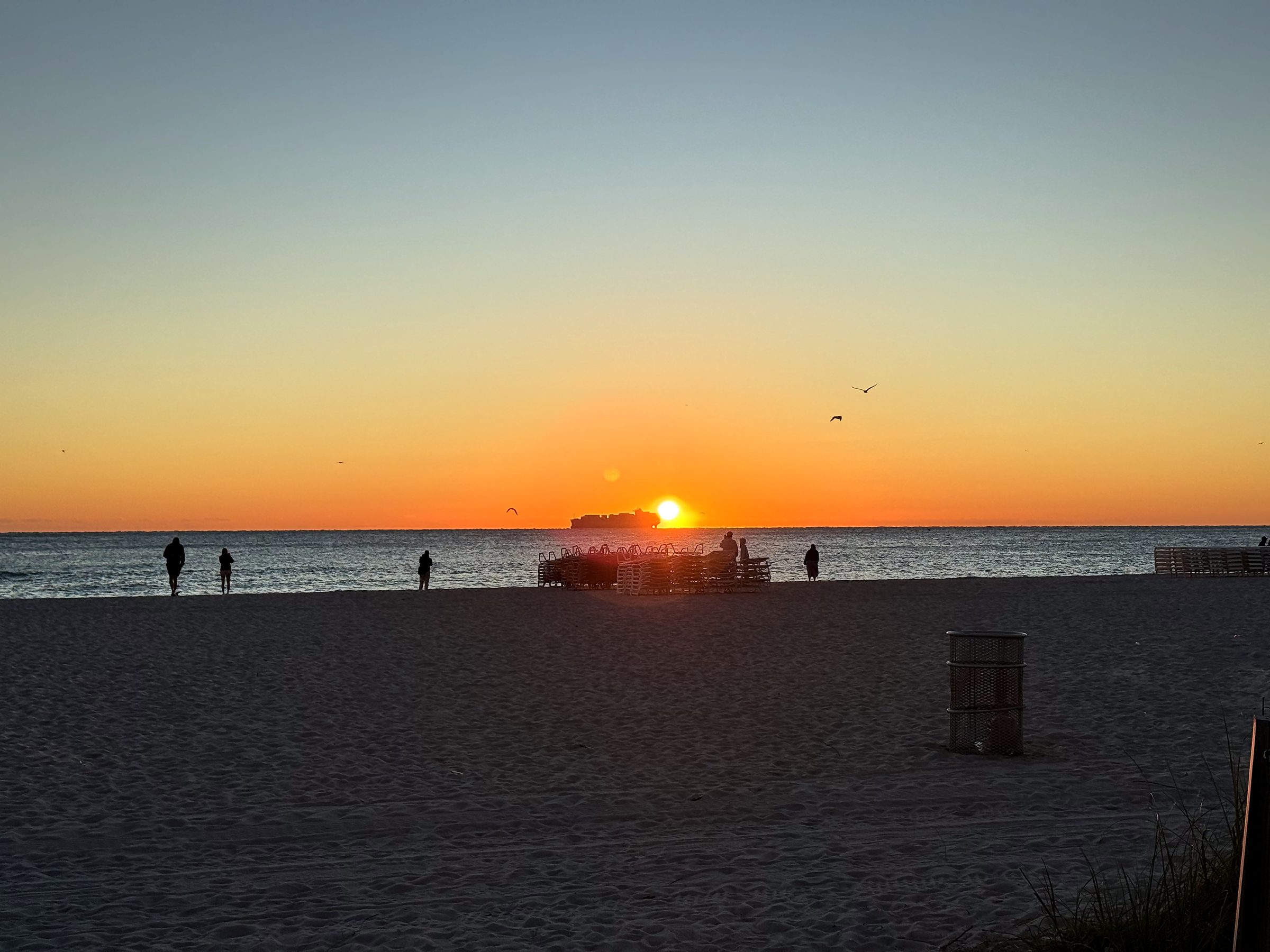 Orange sun near horizon with ship and people silhouetted on beach.