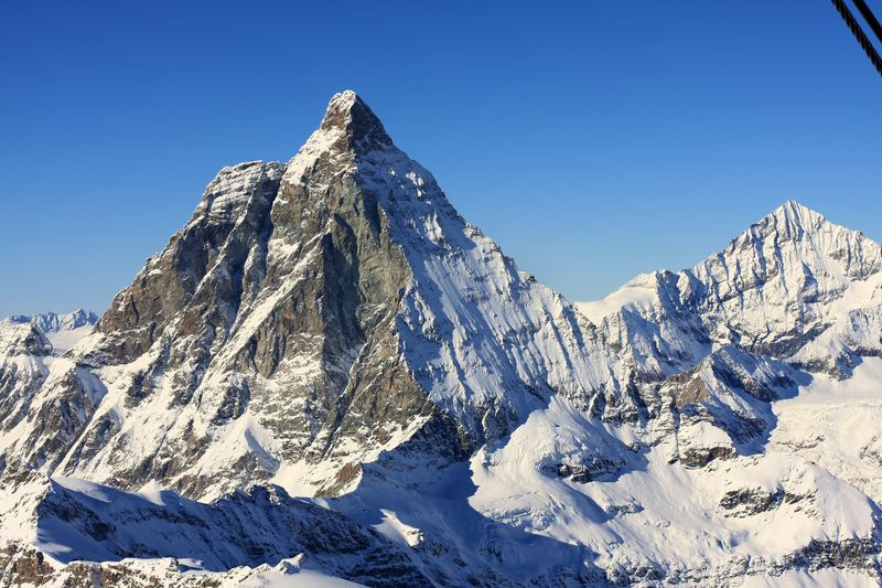 Wide snowy alpine range under clear sky with distant cable-car line.