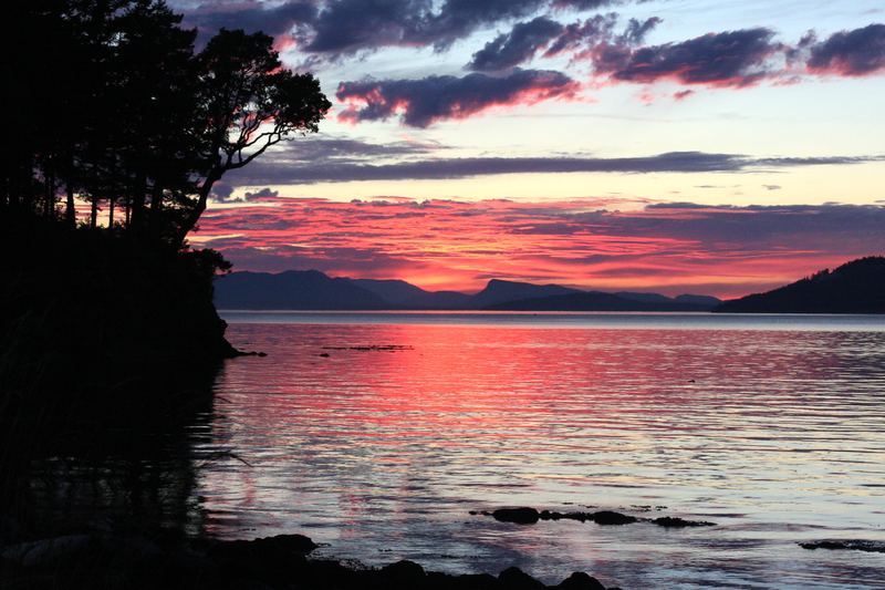 Crimson and purple sunset streaks over water from a dark shoreline.