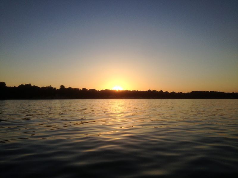Sun centered on the water horizon, treeline eating the day.
