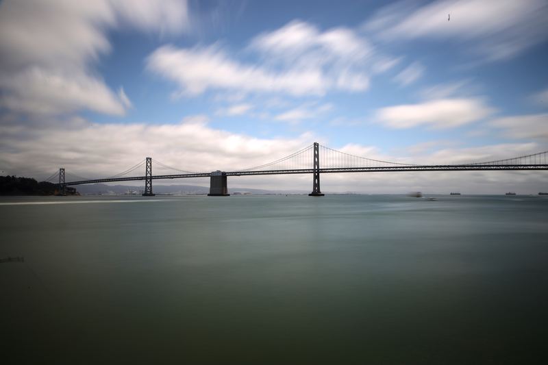 Long bridge across still hazy water under streaked cloudy sky.