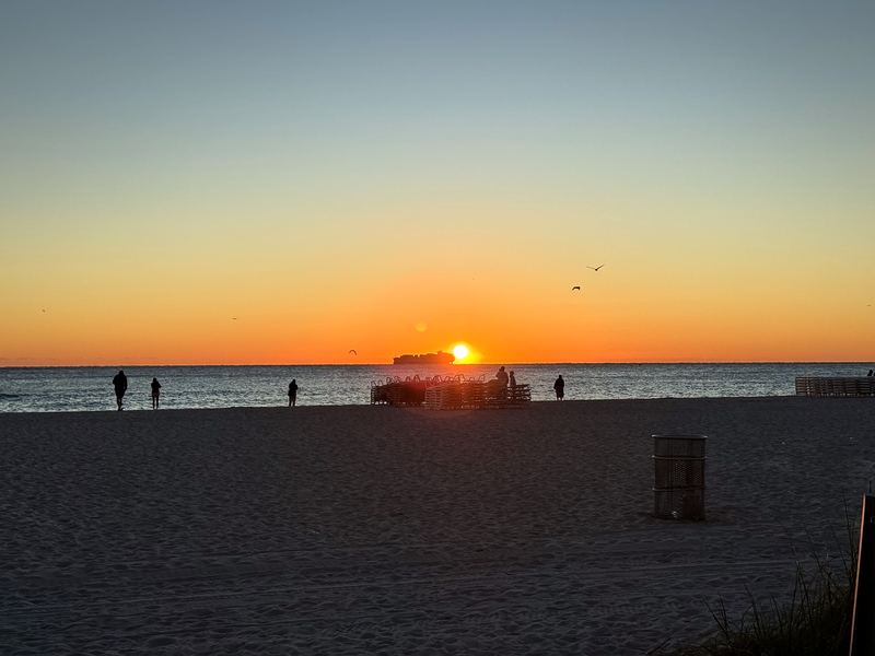 Orange sun near horizon with ship and people silhouetted on beach.